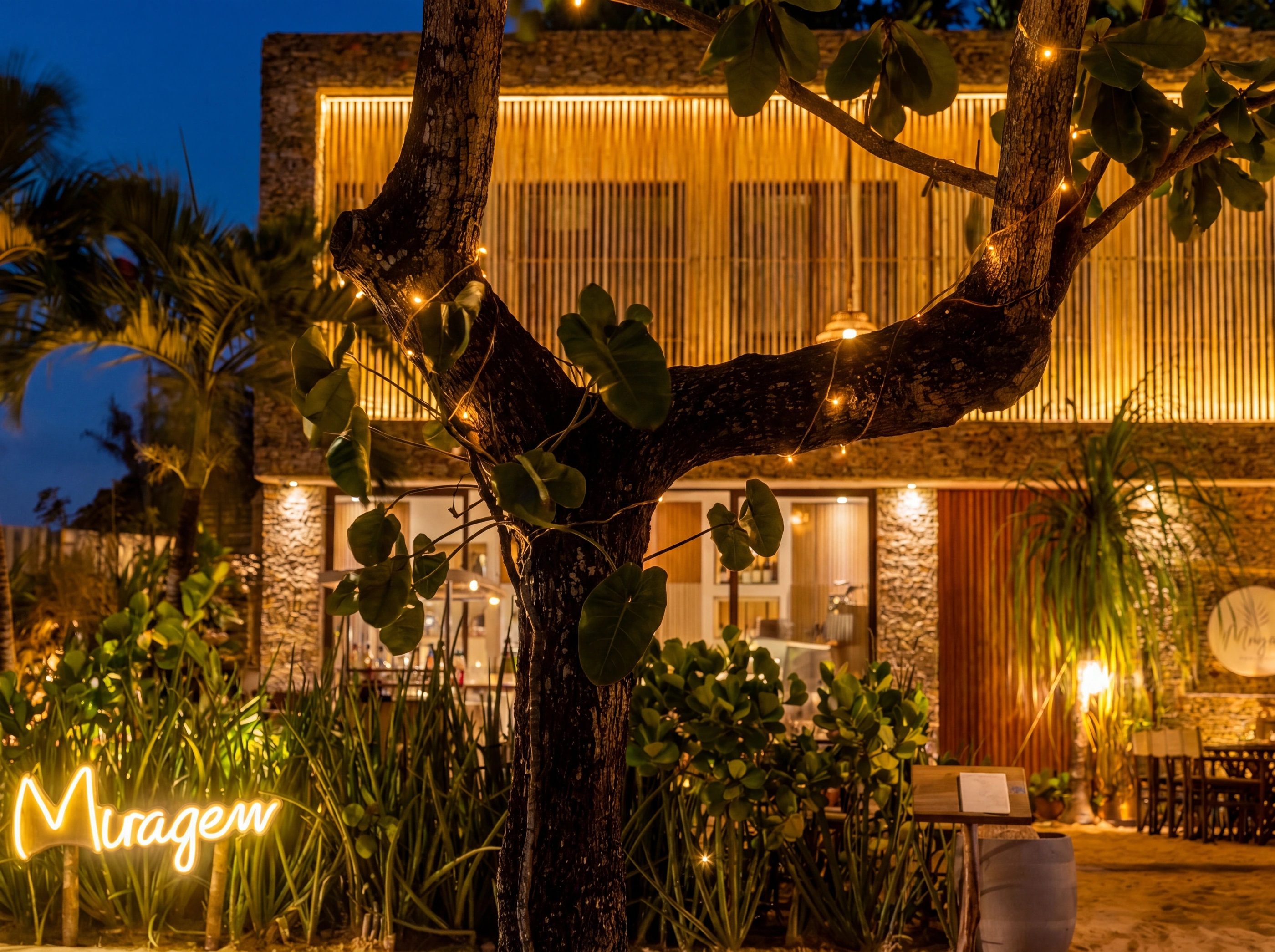 Extérieur du bâtiment Miragem au crépuscule : guirlandes lumineuses le long de la façade en lattes de bois, enseigne au néon Miragem au sol parmi les plantes.