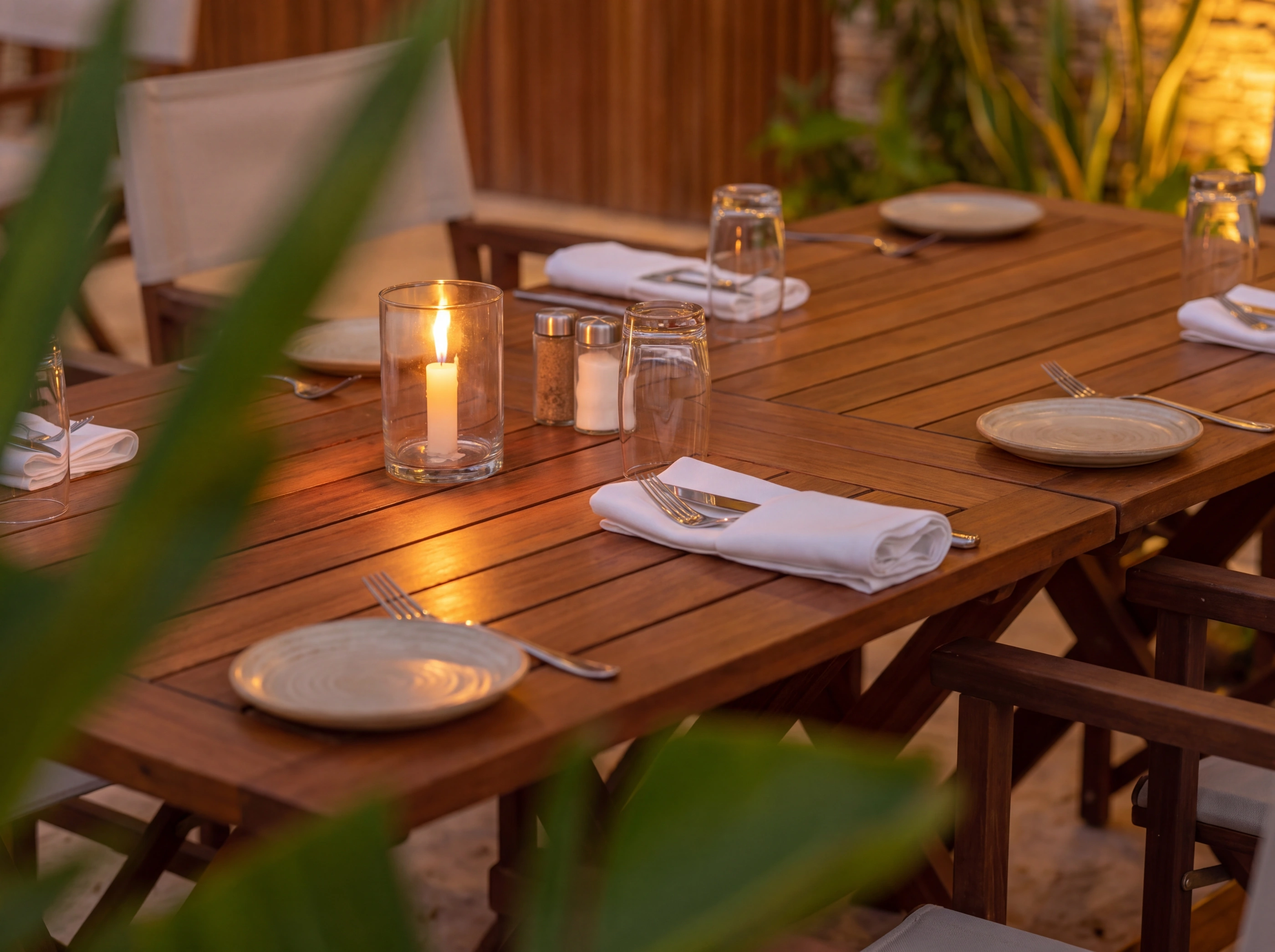 Gros plan d'une table en bois : bougie sous verre, couverts, serviettes en lin, assiettes en céramique, feuillage tropical au premier plan.